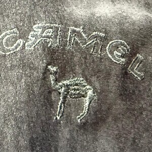 May include: Close-up of a dark gray fabric with the embroidered word "CAMEL" in white thread above a detailed camel silhouette. The fabric has a soft, textured appearance.