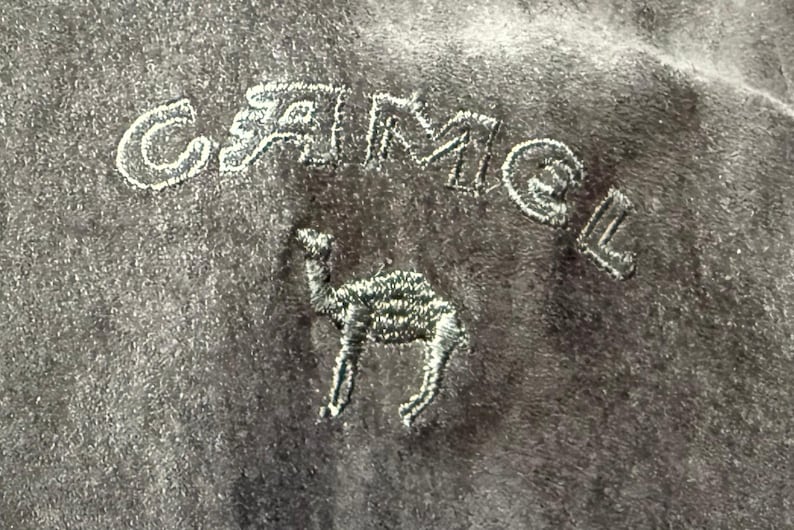 May include: Close-up of a dark gray fabric with the embroidered word "CAMEL" in white thread above a detailed camel silhouette. The fabric has a soft, textured appearance.