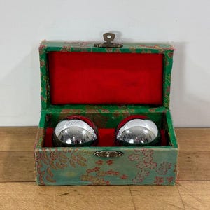 May include: A pair of shiny chrome Baoding balls rest inside an open, ornate green box. The box is lined with red fabric and has a floral pattern. The box is closed with a small metal clasp.