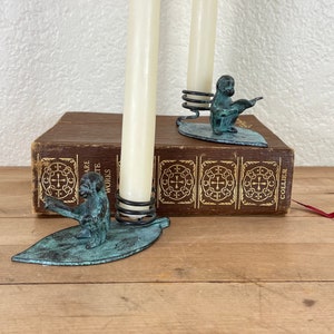 May include: Two antique bronze candle holders. Each features a monkey figure on a leaf-shaped base, with a spiral metal holder for the candle. The candles are tall and ivory-colored. The candle holders are placed on a wooden surface.