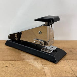 May include: A desktop stapler featuring a polished chrome metal body with a black plastic base and a black top press lever. The stapler has a visible metal mechanism at the front, a silver-colored staple plate, and exposed screws on the side.