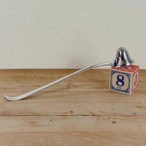 May include: A silver-colored ladle with a long, curved handle rests on a wooden surface. The ladle's bowl is bell-shaped. A red, white, and blue block with the number 8 is next to the ladle. The background is a light beige wall.