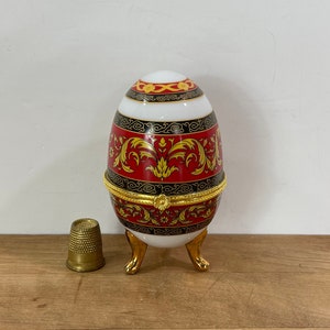 May include: A decorative egg-shaped trinket box with a hinged lid, featuring a red, gold, and black floral design. The egg rests on three gold-colored feet. A small brass thimble is next to the egg.