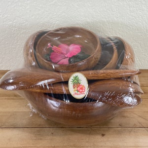 May include: A set of wooden salad bowls and serving spoons, wrapped in clear plastic. The bowls are various sizes, with a large bowl at the bottom. A pink hibiscus flower rests inside one bowl. A sticker with the word "Hawaii" is visible.