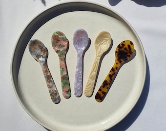 May include: Six colorful spoons with different patterns and designs. The spoons are made of a translucent material and have a rounded shape. The spoons are arranged in a circle on a white plate.