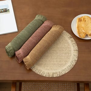 Puede incluir: Tres manteles individuales tejidos con bordes de flecos en verde, rosa y beige. Un mantel individual redondo tejido con bordes de flecos está en primer plano.