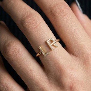 Peut inclure: Une bague dorée ornée des lettres majuscules L et R placées côte à côte. Les lettres sont incrustées de petites pierres claires et scintillantes sur toute leur surface. L'anneau de la bague est fin, lisse et d'aspect métallique.