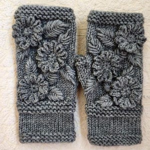May include: A pair of charcoal gray fingerless gloves featuring a textured knit pattern. Each glove is adorned with three-dimensional knitted flowers and leaves across the back, finished with a ribbed cuff at the wrist opening.