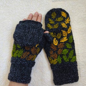 May include: A pair of dark charcoal gray knit convertible mittens featuring a fold-over cap and an embroidered pattern of fern leaves in shades of yellow, lime green, and brown. The knit fabric has a heathered texture throughout.