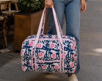 May include: A rectangular duffel bag featuring a dark blue fabric base with a repeating pink and white floral pattern. The bag includes two long pink and white checkered fabric handles and a matching checkered trim along the seams and zipper closure.