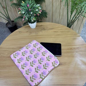 May include: A pink floral pattern zippered pouch with a white zipper, lying on a wooden table next to a black smartphone. The pouch features a repeating pattern of purple flowers and green leaves. Decorative plants are in the background.