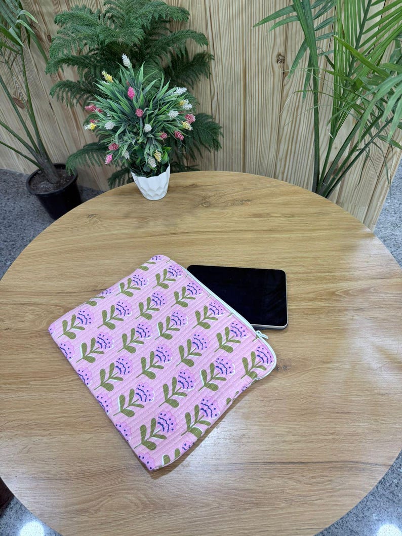 May include: A pink floral pattern zippered pouch with a white zipper, lying on a wooden table next to a black smartphone. The pouch features a repeating pattern of purple flowers and green leaves. Decorative plants are in the background.