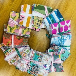 May include: A circular arrangement of colorful fabric squares, each bundle tied with a ribbon. The fabrics display various patterns, including floral, striped, and animal prints, in shades of pink, green, blue, and orange. The arrangement is set on a wooden surface.