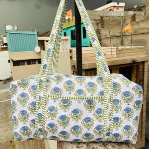 May include: A white tote bag with a blue floral pattern and green trim. The bag has two long handles with a green and white striped design. The bag is made of a quilted fabric and is sitting on a stone surface.