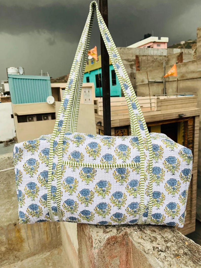 May include: A white tote bag with a blue floral pattern and green trim. The bag has two long handles with a green and white striped design. The bag is made of a quilted fabric and is sitting on a stone surface.