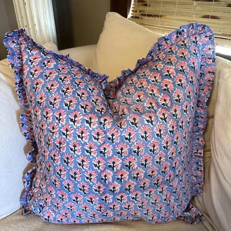 May include: A square throw pillow featuring a light blue base with a repeating pattern of pink flowers and dark green stems. The pillow is finished with a ruffled fabric border along all four edges, creating a decorative frame around the floral print.