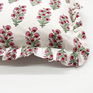 May include: A white rectangular throw pillow featuring a repeating floral pattern of pink flowers with green stems and leaves. The pillow is finished with a gathered fabric ruffle trim along the edge, which also displays the same floral print.
