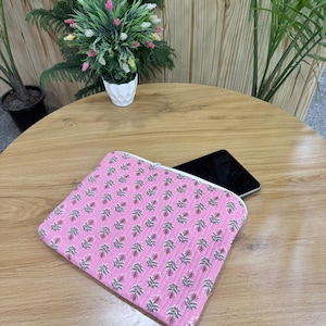 May include: A pink quilted tablet sleeve with a floral pattern in red, green, and white. The sleeve is rectangular with a white zipper. A black tablet is partially visible inside the sleeve. The sleeve is on a wooden table.