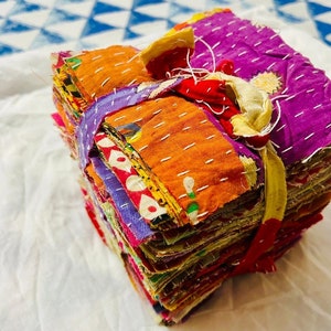 May include: A stack of square fabric swatches featuring various vibrant colors including purple, orange, and red, held together by a tied fabric strip. Each piece of cloth displays visible white running stitch embroidery patterns throughout.