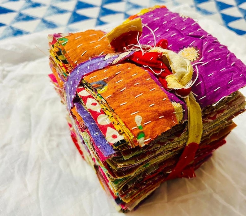 May include: A stack of square fabric swatches featuring various vibrant colors including purple, orange, and red, held together by a tied fabric strip. Each piece of cloth displays visible white running stitch embroidery patterns throughout.