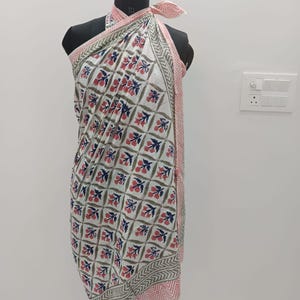 May include: A white sarong with a repeating floral pattern in red, blue, and green. The edges are trimmed with a pink and gray geometric design. The sarong is draped over a mannequin.