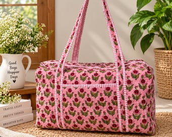 May include: A quilted duffel bag in a light pink fabric featuring a repeating dark red floral pattern with green leaves. The bag includes two long handles and trim accents in a white and pink striped fabric, with a rectangular shape and stitching.
