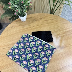 May include: A navy blue zippered pouch with a floral pattern of pink and white flowers and green leaves. The pouch is on a wooden table next to a black tablet. A small potted plant is in the background.