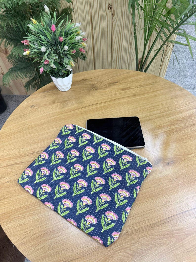 May include: A navy blue zippered pouch with a floral pattern of pink and white flowers and green leaves. The pouch is on a wooden table next to a black tablet. A small potted plant is in the background.