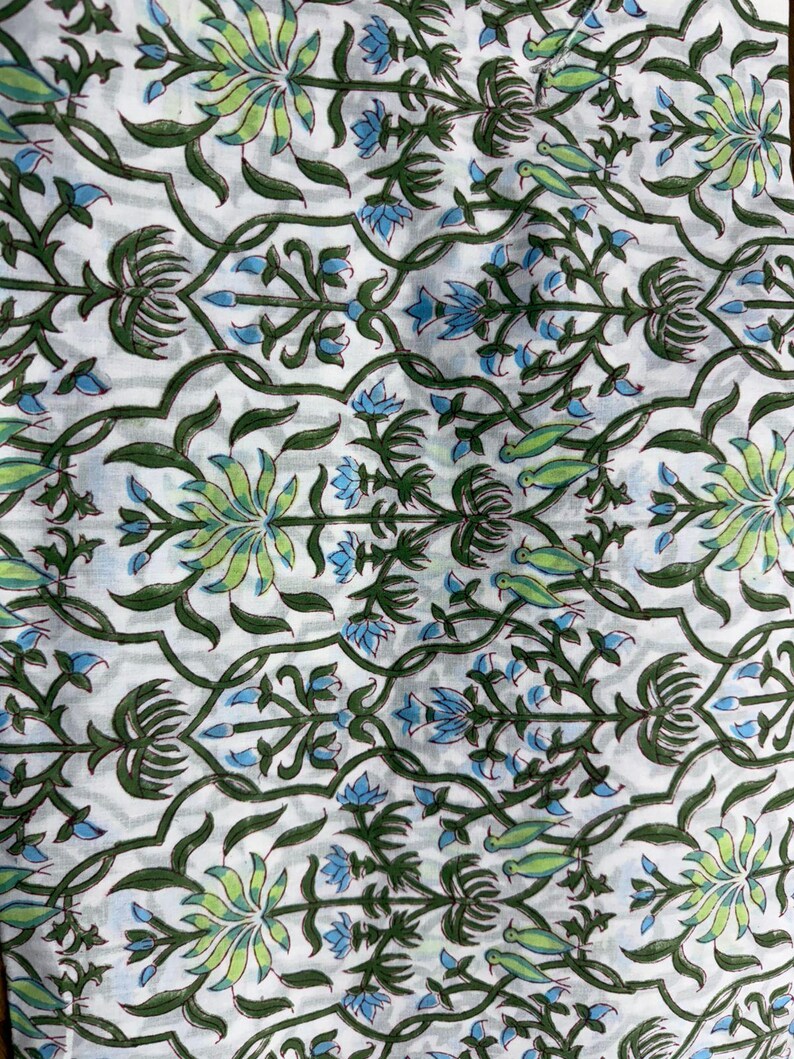 Pu&ograve; includere: Tessuto bianco con un motivo floreale ripetuto in verde, verde chiaro e blu. Il design include fiori stilizzati, foglie e forme simili a uccelli, creando un tema botanico. Il tessuto sembra essere cotone o un materiale leggero simile.