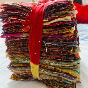 May include: A tall stack of square fabric scraps featuring various colors and patterns, bound together by a red and yellow fabric strip. Each piece of fabric displays visible white running stitch embroidery details along the edges and surfaces.