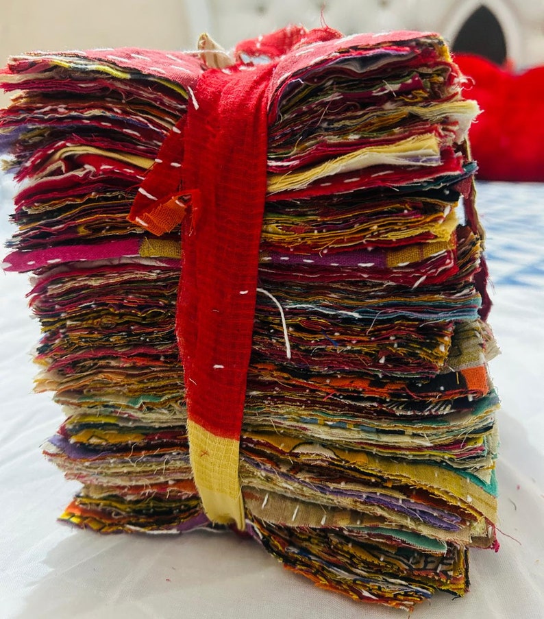 May include: A tall stack of square fabric scraps featuring various colors and patterns, bound together by a red and yellow fabric strip. Each piece of fabric displays visible white running stitch embroidery details along the edges and surfaces.