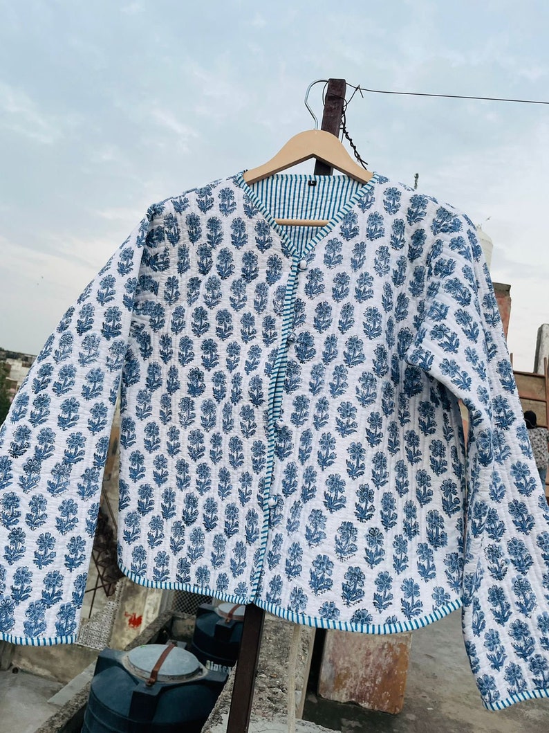 Pu&ograve; includere: Una giacca trapuntata bianca con stampa floreale blu e fodera a righe blu e bianche. La giacca ha una chiusura con bottone e maniche lunghe.