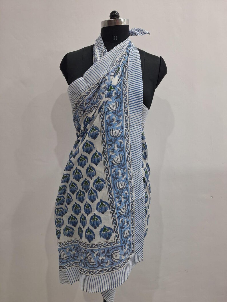 May include: A white and blue floral print sarong draped on a mannequin. The sarong features a repeating pattern of blue flowers and a striped border. The fabric is lightweight and appears to be cotton.
