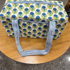 May include: A rectangular tote bag featuring a floral design in blue, yellow, and green on a white backdrop. It has blue and white striped handles and trim. The bag is placed on a wooden table.