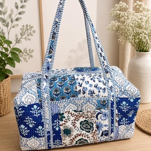 May include: A rectangular quilted tote bag featuring a patchwork design of various blue and white floral and paisley patterns. The bag has two long shoulder straps with a blue and white striped pattern and a zippered top closure.