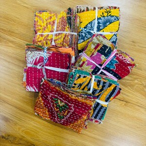 May include: Stacks of colorful, patterned fabric squares tied with white string. The fabrics feature various designs in red, yellow, blue, and green. The squares are arranged on a light brown wooden surface.