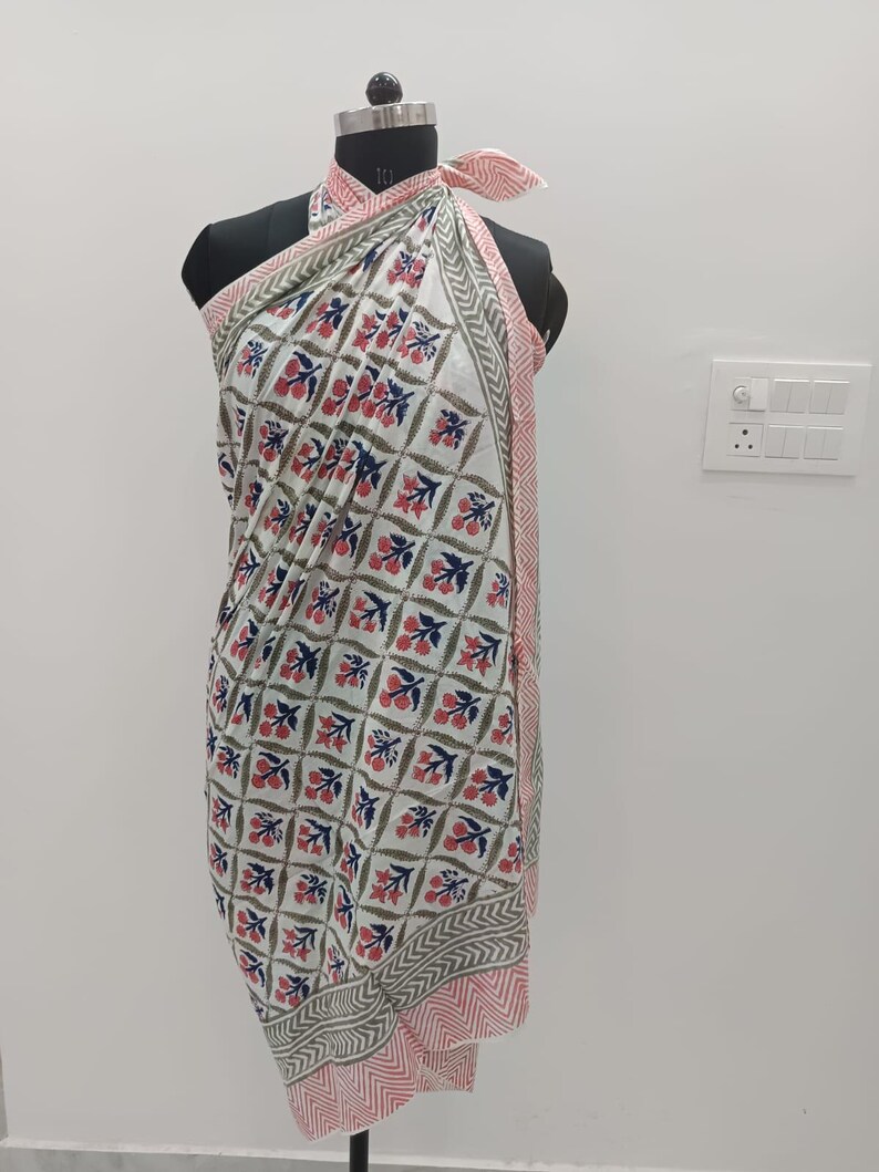 May include: A white sarong with a repeating floral pattern in red, blue, and green. The edges are trimmed with a pink and gray geometric design. The sarong is draped over a mannequin.
