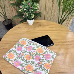 May include: A white floral laptop sleeve featuring a pattern of pink and orange flowers with green leaves. The sleeve is on a wooden table next to a black tablet. A small white pot with a plant is in the background.