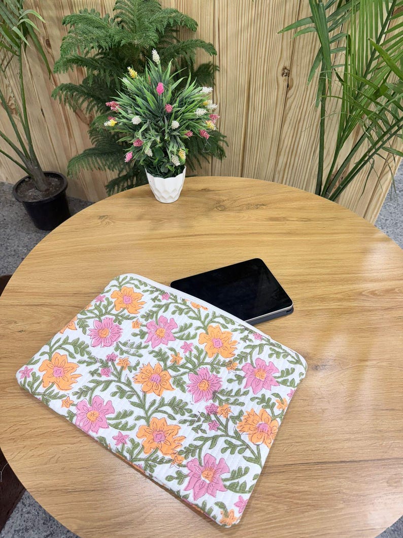 May include: A white floral laptop sleeve featuring a pattern of pink and orange flowers with green leaves. The sleeve is on a wooden table next to a black tablet. A small white pot with a plant is in the background.