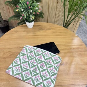 May include: A white and pink floral patterned laptop sleeve with a green leaf design. The sleeve is on a wooden table with a black tablet. A small white pot with a plant is in the background.