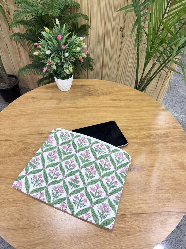 May include: A white and pink floral patterned laptop sleeve with a green leaf design. The sleeve is on a wooden table with a black tablet. A small white pot with a plant is in the background.