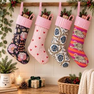 Puede incluir: Cuatro calcetines de Navidad colgantes con puños de rayas rosas y blancas. El primero es negro con patrones florales rosas y naranjas, el segundo es rosa claro con corazones rojos, el tercero es blanco con enredaderas florales azules y el cuarto tiene rayas naranjas con ilustraciones de tigres.