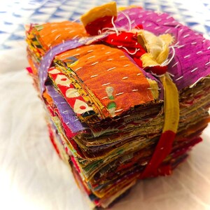 May include: A square stack of assorted fabric scraps featuring various colors and patterns, including orange, purple, and red. The fabric layers are held together by a yellow fabric tie and feature visible white running stitch embroidery throughout.