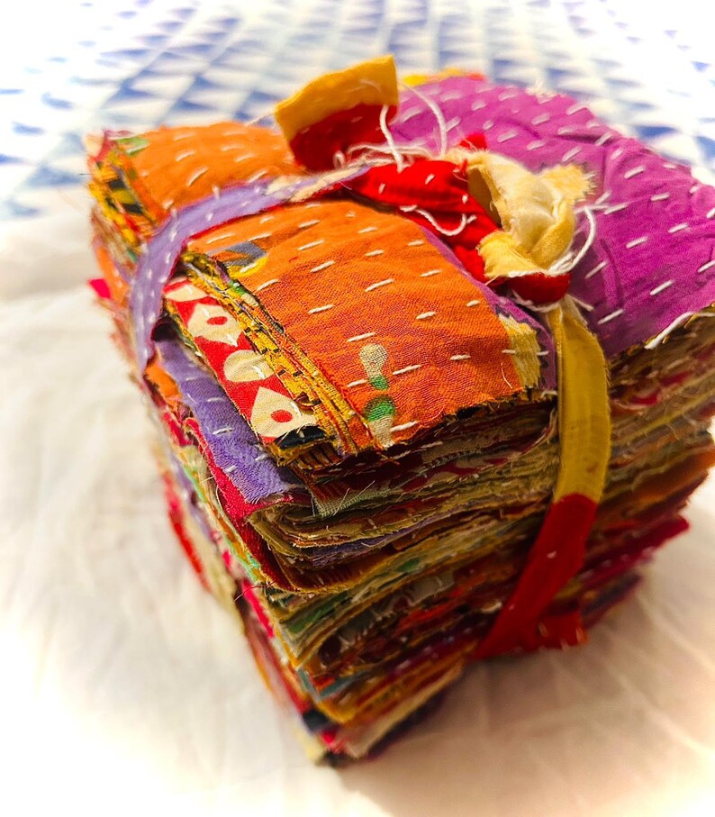 May include: A square stack of assorted fabric scraps featuring various colors and patterns, including orange, purple, and red. The fabric layers are held together by a yellow fabric tie and feature visible white running stitch embroidery throughout.