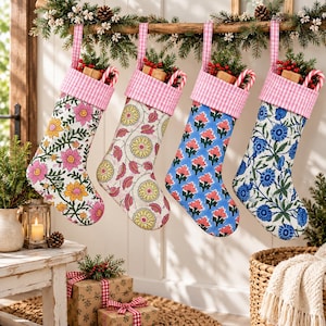Könnte beinhalten: Vier Weihnachtsstrümpfe hängen in einer Reihe, jeder mit einem rosa-weiß karierten Umschlag. Die Strümpfe zeigen verschiedene Blumenmuster in Rosa, Gelb und Blau, wobei kleine Geschenkboxen und rote Beeren oben aus den Strümpfen herausragen.