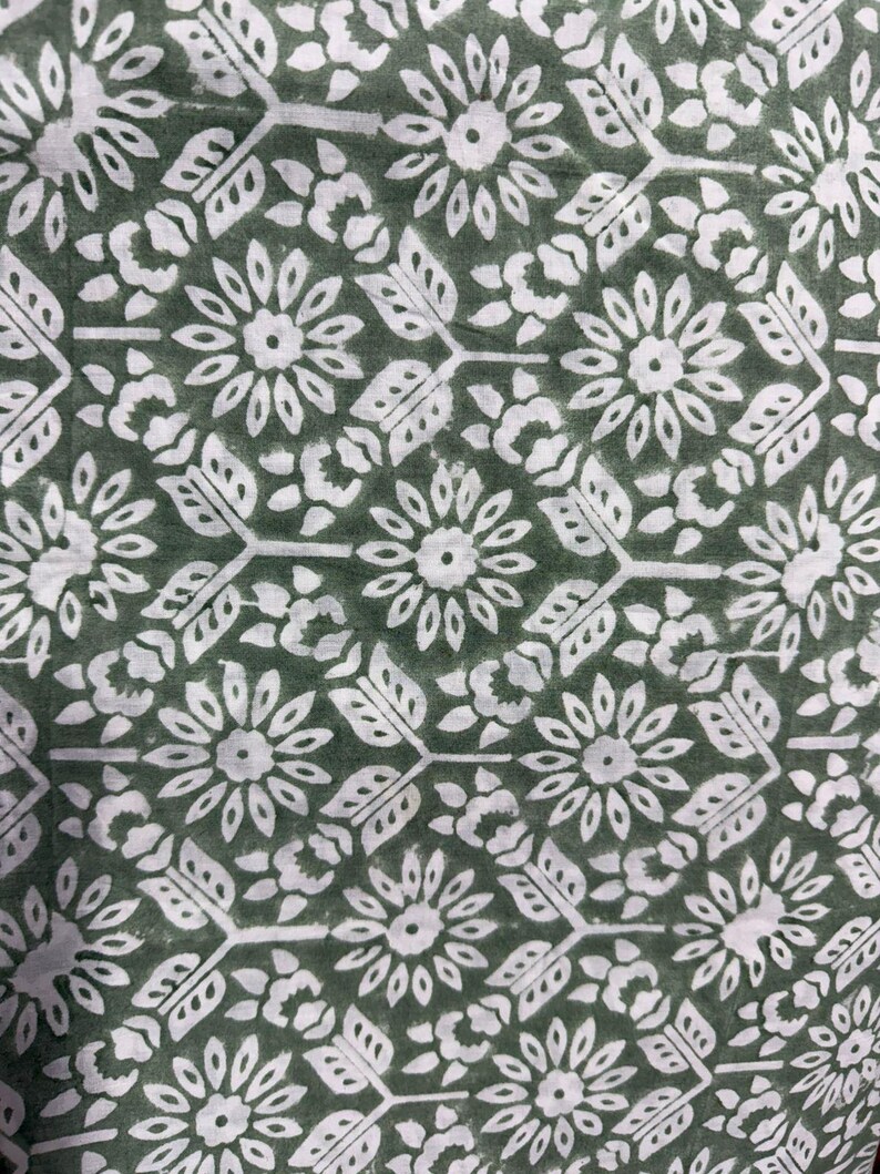 Pu&ograve; includere: Primo piano di un tessuto con un motivo floreale ripetuto. Il design presenta fiori bianchi, foglie e forme geometriche su uno sfondo verde scuro. Il motivo &egrave; uniformemente distanziato e dettagliato.
