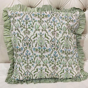 May include: A square throw pillow featuring a white base with a repeating pattern of green vines, light blue flowers, and small green birds. The pillow is finished with a ruffled border made of fabric with thin green and white vertical stripes.