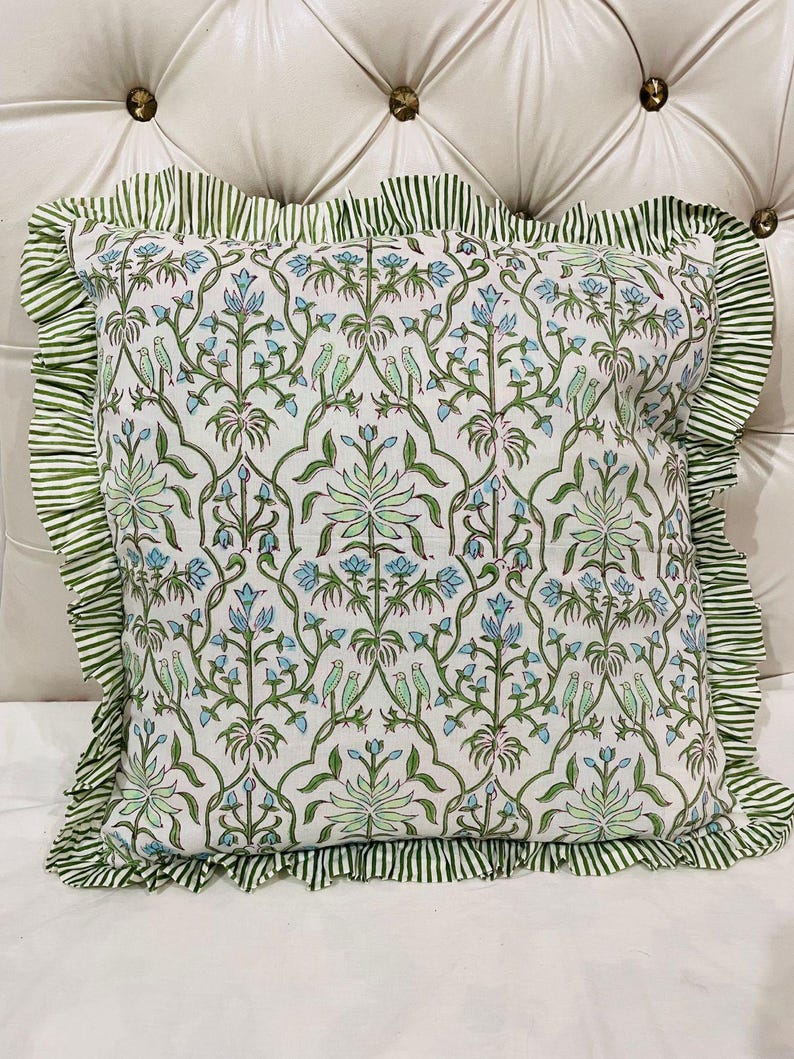 May include: A square throw pillow featuring a white base with a repeating pattern of green vines, light blue flowers, and small green birds. The pillow is finished with a ruffled border made of fabric with thin green and white vertical stripes.