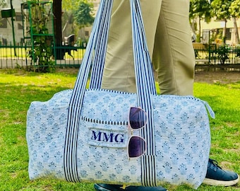 May include: A blue and white floral print duffel bag with navy blue and white striped handles. The bag has a small pocket with the initials "MMG" and a pair of sunglasses attached. The bag is held by a person standing on grass.