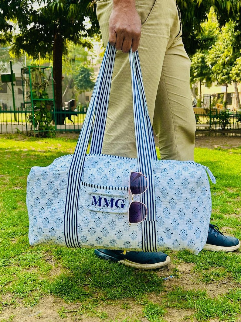 May include: A blue and white floral print duffel bag with navy blue and white striped handles. The bag has a small pocket with the initials "MMG" and a pair of sunglasses attached. The bag is held by a person standing on grass.